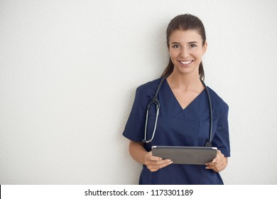 Young woman doctor or nurse in blue scrubs holding a tablet pc standing in front of a white wall