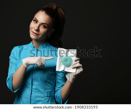 Young woman doctor nurse in blue uniform and protective latex gloves shows points finger at special medical tool ring over dark background with copy space. Medicine workers and healthcare concept