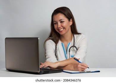 Young Woman Doctor Sitting At Office Desk And Working On Laptop. Online Doctor Concept. Portrait Of A Nurse  On White Background, Close-up