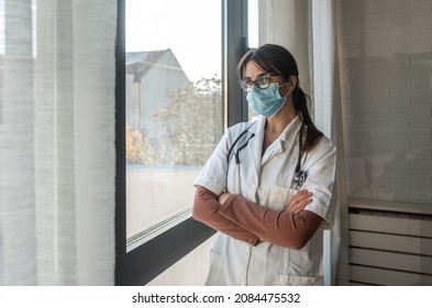 Young Woman Doctor Standing Tired And Exhausted Near Hospital Window. Female Health Care Medical Worker With Protective Facial Mask Taking A Break From Work At The Clinic Feeling Overworked.