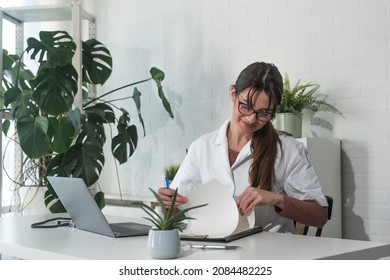 Young Woman Doctor Starting Her Day Shift At The Hospital Office. Female Health Care Medical Worker Arranging Her Desk At Clinic With Laptop Paper And Stethoscope Before First Patient Enter.