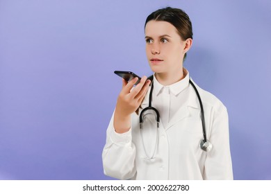 Young Woman Doctor Talk To Smartphone For Order Command, Posing Isolated Over Violet Studio Background With Copy Space. Voice Recognition Biometric, Speech Detect In Healthcare Technology Concept