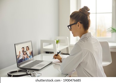 Young Woman Doctor Therapist In White Uniform Listening To Complaints Of Happy Family With Child During Online Consultation On Laptop From Medical Clinic Office. Telehealth, Telemedicine Cocnept