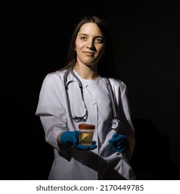 A Young Woman Doctor In A White Coat And Medical Gloves Holds A Jar With Urine Tests In Her Hand On A Black Background