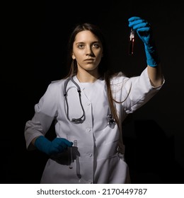 Young Woman Doctor In A White Coat And Medical Gloves Holds Test Tubes With Blood In Her Hands On A Black Background