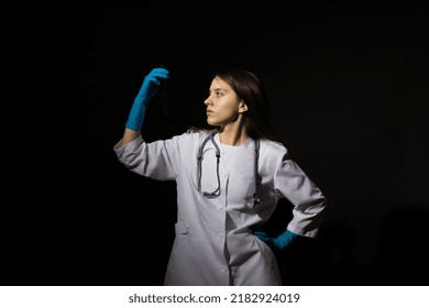 Young Woman Doctor In A White Coat And Medical Gloves Holds Test Tubes With Blood In Her Hands On A Black Background