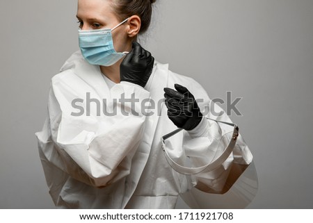 young woman doctor in white protective suit and black gloves removes medical mask from her face on grey background.