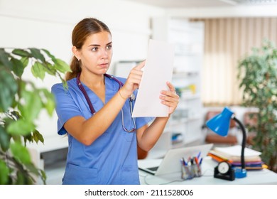 Young Woman Doctor Working In The Clinic Is Attentive Studying The Patient's Outpatient Card, Standing In The Resident's ..office. Close-up Portrait..