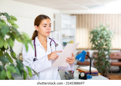 Young Woman Doctor Working In The Clinic Is Attentive Studying The Patient's Outpatient Card, Standing In The Resident's ..office. Close-up Portrait..