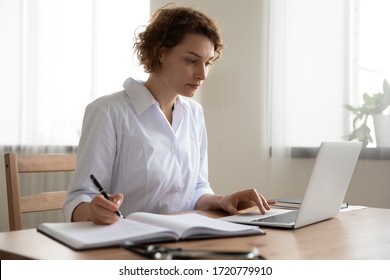 Young woman doctor working on laptop taking notes at workplace. Female physician writing in notebook using computer sitting at desk. Professional medic therapist everyday routine in hospital office. - Powered by Shutterstock