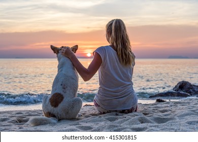 Young woman with dog sitting on the beach and watching the sunset
