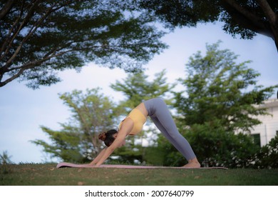 A Young Woman Doing Downward Facing Dog Yoga Pose On Grass Floor With Mat Among Big Tree At Frontyard With Peaceful And Happiness.