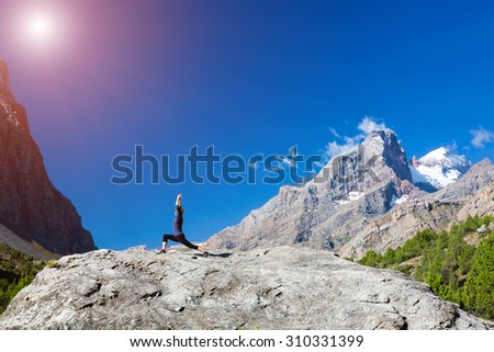 Young woman doing exercise.
Cute Girl Doing Yoga Fitness to Stretch Body Staying on High Rock at Mountain Panoramic Landscape Outdoor Sunny Sky Peaks Wilderness Country Smiling Beautiful Face Sunshine