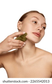 Young woman doing facial massage with guasha scrapper. Concept of beauty, skin care, self-massage, spa procedure at home. Model without makeup and well-kept skin