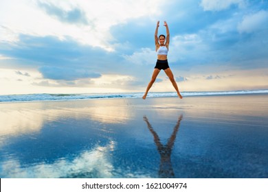 Young woman doing jumping jack or star jumps exercise to burn fat, keep fit. Sunset beach, blue sky background. Healthy lifestyle at training camp, outdoor fitness activity, family summer holiday.
