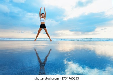 Young Woman Doing Jumping Jack Or Star Jumps Exercise To Burn Fat, Keep Fit. Sunset Beach, Blue Sky Background. Healthy Lifestyle At Training Camp, Outdoor Fitness Activity, Family Summer Holiday.