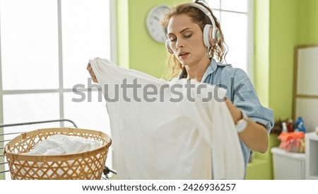 Young woman doing laundry indoors, folding clothes with headphones on in a brightly lit room.