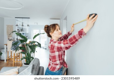 Young woman doing measuring with a measure tape on the wall. Girl wants to put a picture on the wall at home. Housekeeping work. Doing repair herself. DIY, gender equality in work concept. - Powered by Shutterstock