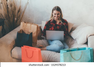 Young woman doing online shopping at home using laptop. Look at its screen and smile. She sits on sofa surrounded with shopping bags. Happy satisfied customer with purchase Foto Stock