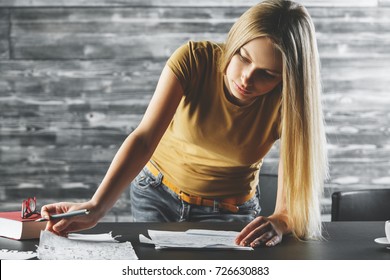 Young woman doing paperwork at office desk with book, coffee cup and other items. Blurry wooden wall in the background. Project, contract, document concept 