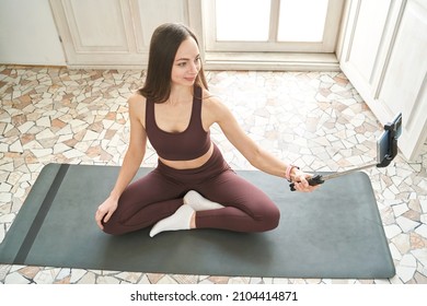 Young Woman Doing Self Portrait On Yoga Mat. Home Vacation Indoors Portrait. Blogger Selfie Stick. Alone In Interior. Happy Emotion. Smiling Female Person. Lifestyle Action. Posing Influencer