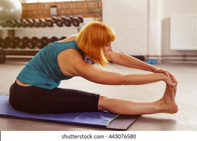 Young Woman Doing Stretching Exercises In A Gym Sitting On A Mat Stretching Forwards To Touch Her Toes, Close Up Side View