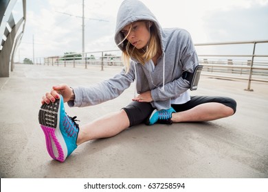 Young woman doing stretching on a modern bridge after intensive sports training. She is blonde, wearing hoodie and listening music - Powered by Shutterstock