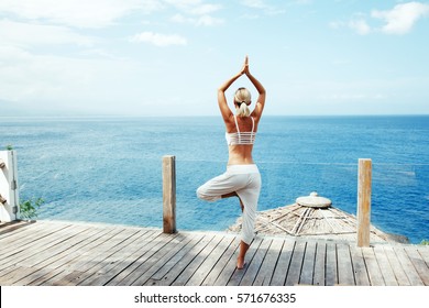 Young woman doing yoga asana in the nature with the ocean view. Morning workout outdoors, sports and healthy lifestyle.