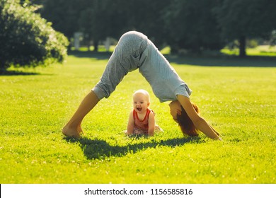 Young woman doing yoga with baby in the summer park. Beautiful mother and daughter like a little buddha exercising on green grass on nature. Family outdoors. Parent with child spend time together.