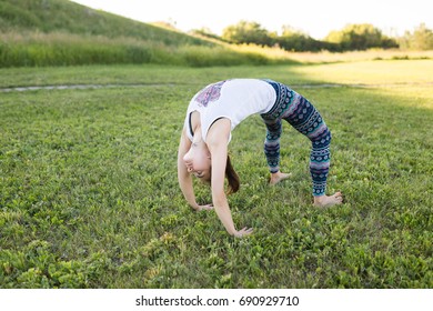 Young woman doing yoga Bridge pose. Asana Chakrasana or Dhanurasana. Beautiful indian woman enjoying yoga outdoors in sport clothes, working out in park with green grass