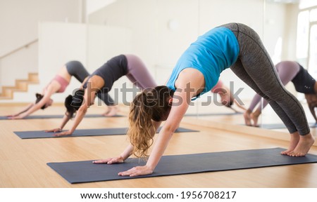 Young woman doing yoga with group in fitness studio, standing in stretching asana Adho Mukha Shvanasana known as Downward Dog pose