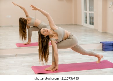 Young woman doing yoga in the gym. A girl with long hair and in a beige tracksuit stands in a parivritta trikonasana pose on a pink carpet. A woman performs parivritta trikonasana.