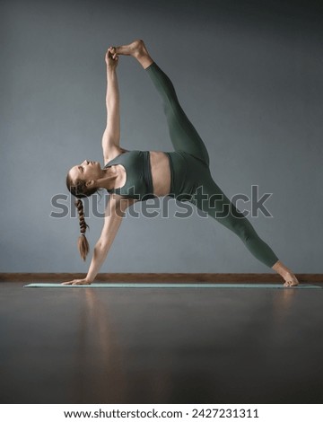Young woman doing yoga in gym on mat. Utthita Vasisthasana. Side plank pose. Yoga-coach. touching toes with hand in side plank. balance. Stand on one arm and one leg. Gray background. Front view. 