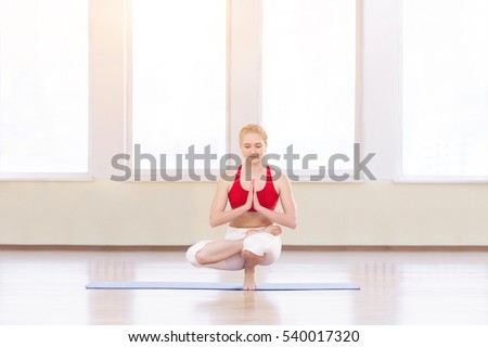 Young woman doing yoga pose half Lotus toe balance. Asana Utkatasana. Beautiful blond woman enjoying yoga indoors in sport clothes, working out in gym class.