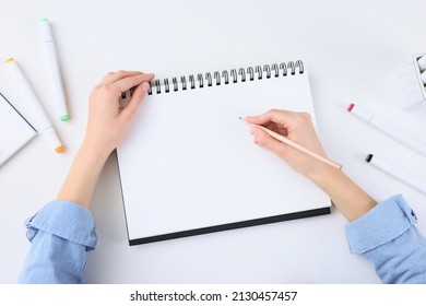 Young woman drawing in sketchbook at white table, top view