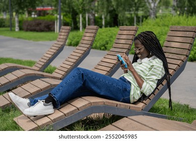 Young woman with dreadlocks smiles while using her smartphone on a park bench, enjoying a sunny day - Powered by Shutterstock