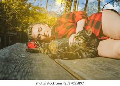 Young woman with dreadlocks wearing red and black lumberjack shirt sleeping with tabby cat on wooden table in mountain forest during sunny autumn day - Powered by Shutterstock