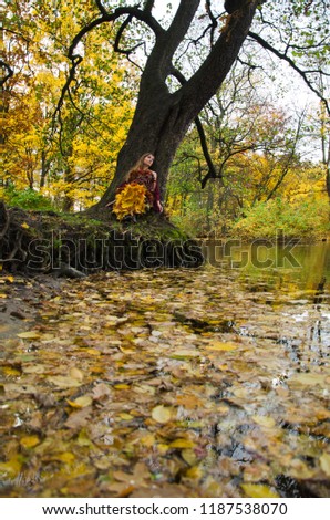 young woman in a dress of autumn leaves and a Burgundy shawl in the woods near the pond sitting by an old tree