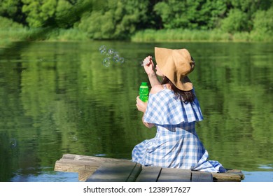 Young Woman In A Dress And A Straw Hat Blowing Bubbles Against T