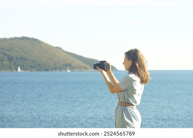 A young woman in a dress takes a photo with a DSLR camera while standing on the seashore. A female tourist takes a photo of the view of the Bay of Kotor from the embankment of the city of Herceg Novi - Powered by Shutterstock