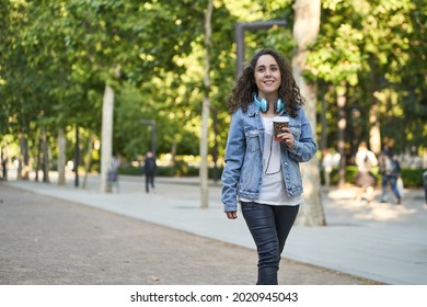 young woman dressed in black leather pants and denim jacket drinking coffee with headphones on her neck