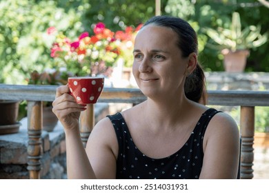 A young woman, dressed in black with white polka dots, enjoys a warm beverage from a red polka dot mug while sitting on a porch in the countryside. - Powered by Shutterstock