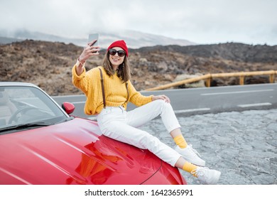 Young woman dressed in bright sweater and hat enjoying road trip on the desert valley, photographing on phone while sitting on the car hood - Powered by Shutterstock