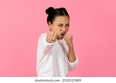 A young woman dressed in casual attire throws a powerful punch while standing confidently in front of a vibrant pink background. Her determined expression showcases her strength and focus. - Powered by Shutterstock