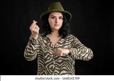 Young Woman Dressed Casually In An Animal Print Shirt, With Black Hair Wearing A Hat Showing Time Is Money Posing On A Black Isolated Backround.