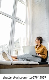 Junge Frau gekleidet, die beim Sitzen auf dem Fensterbrett zu Hause spielend am Laptop arbeitet. Arbeiten Sie von zu Hause aus gemütliche Atmosphäre Konzept