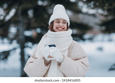 Young woman dressed coat scarf hat and mittens smiles and enjoys hot coffee at snowy winter park