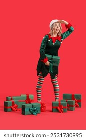 Young woman dressed as elf with Christmas gifts looking somewhere on red background