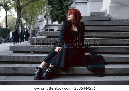 Young woman dressed gothic style in long black dress and cloak wearing red hair sitting alone outdoors in city on the steps of medieval European building