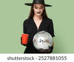 Young woman dressed for Halloween as witch with drink and disco ball on green background